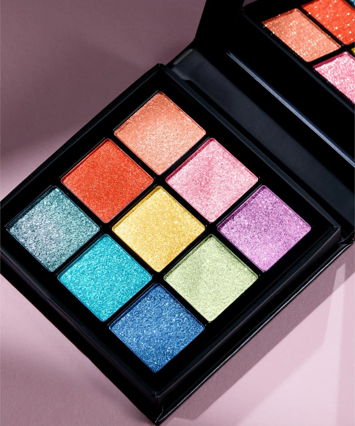 Angled close-up of the open palette on a pink backdrop, shimmering coral, pink, yellow, lilac, aqua and blue pans