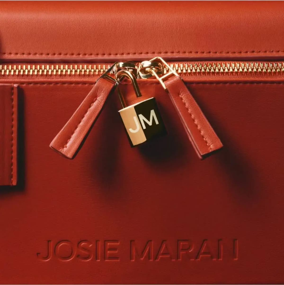 Close-up of gold zipper pulls and a JM padlock on red leather above the embossed Josie Maran logo