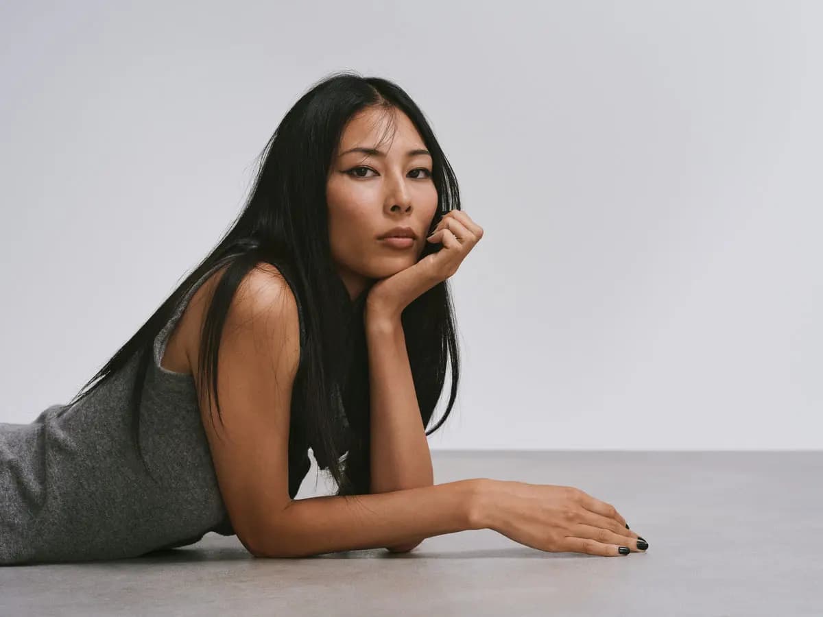 Model with sleek straight black hair lying on a grey floor, chin resting on hand, wearing a grey knit dress