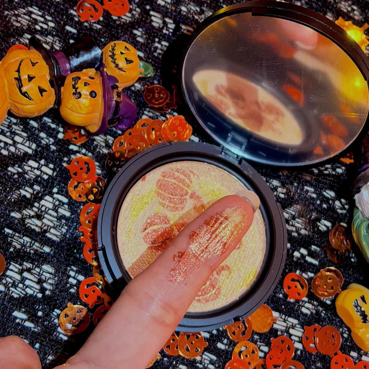 Fingertip swatch of coppery-orange shimmer beside the open pumpkin highlighter and mirror on black lace