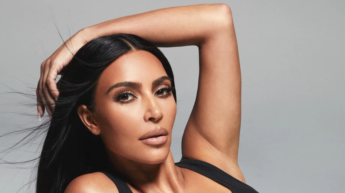 Kim Kardashian posing with one arm raised above her head, wearing a black tank top with glowing bronzed makeup.