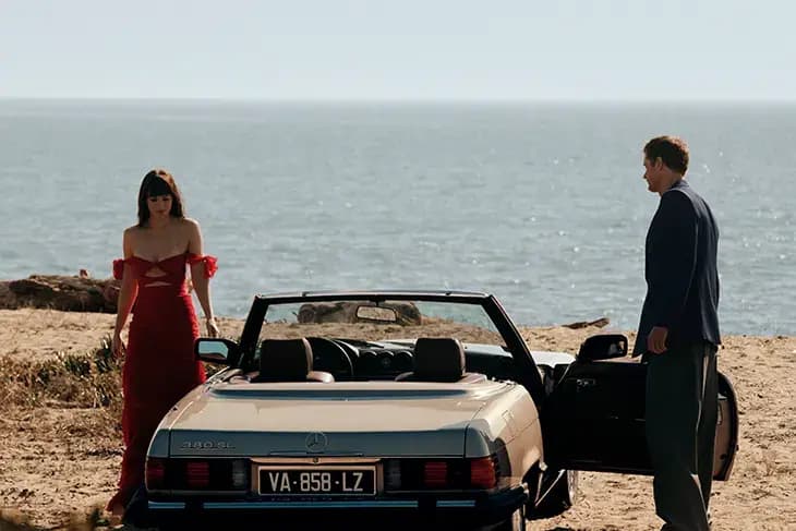 Dakota Johnson in a red dress and Alexander Skarsgård in a navy suit beside a vintage convertible on a seaside cliff in the Vendetta campaign