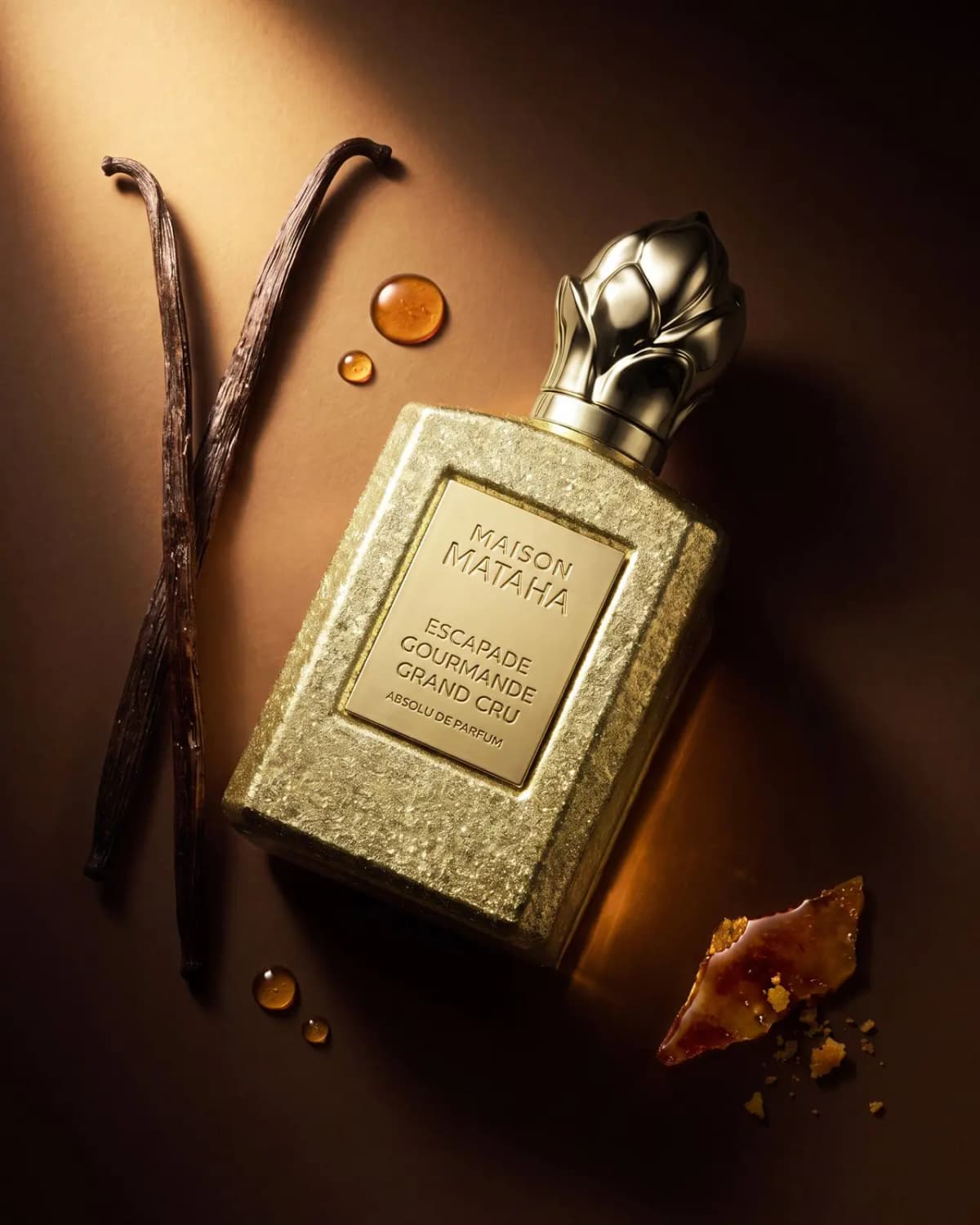Gold-leaf bottle lying diagonally on brown surface with crossed vanilla pods, amber droplets and burnt caramel shard