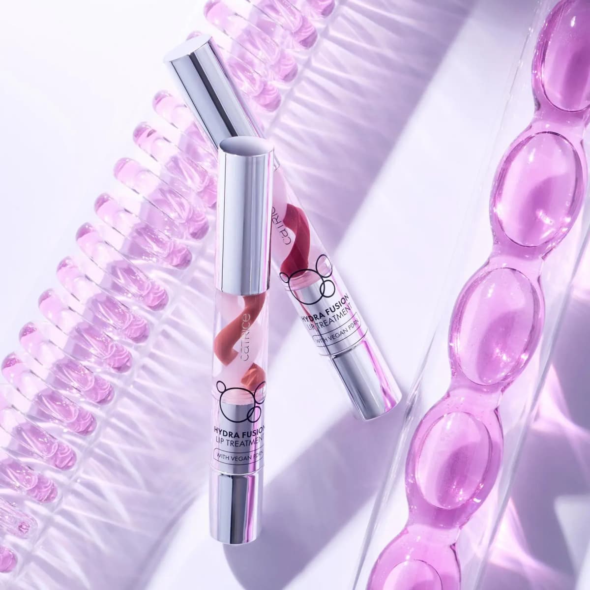 Two Catrice Hydra Fusion Lip Treatment tubes among pink gel capsules and a beaded tube in lilac light