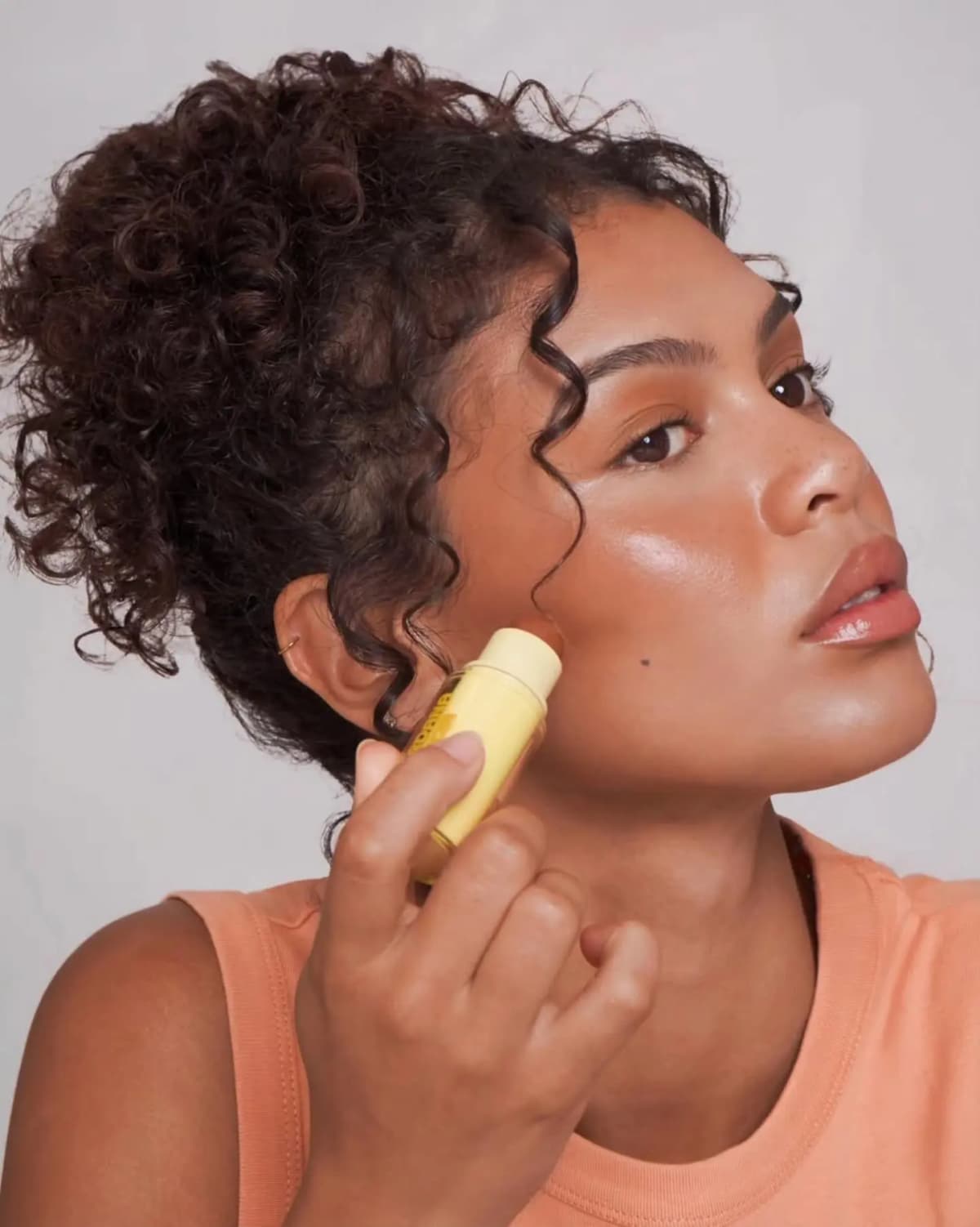 Curly-haired model in an orange tank top applies the cream bronzer stick to her cheekbone