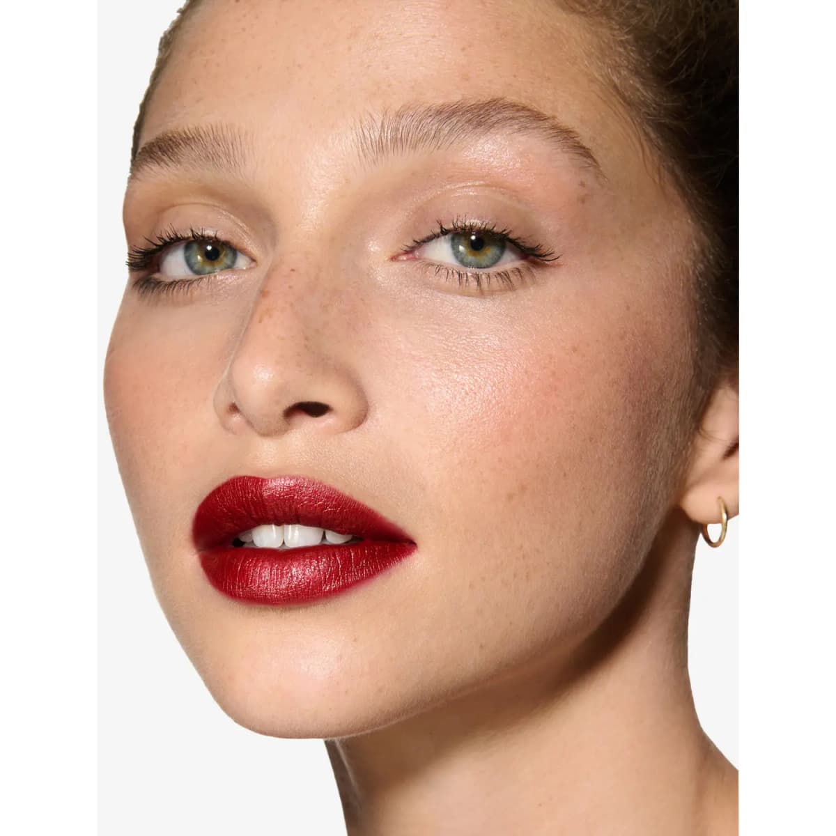 Freckled model with green eyes wearing a metallic deep red lipstick and a small gold hoop earring