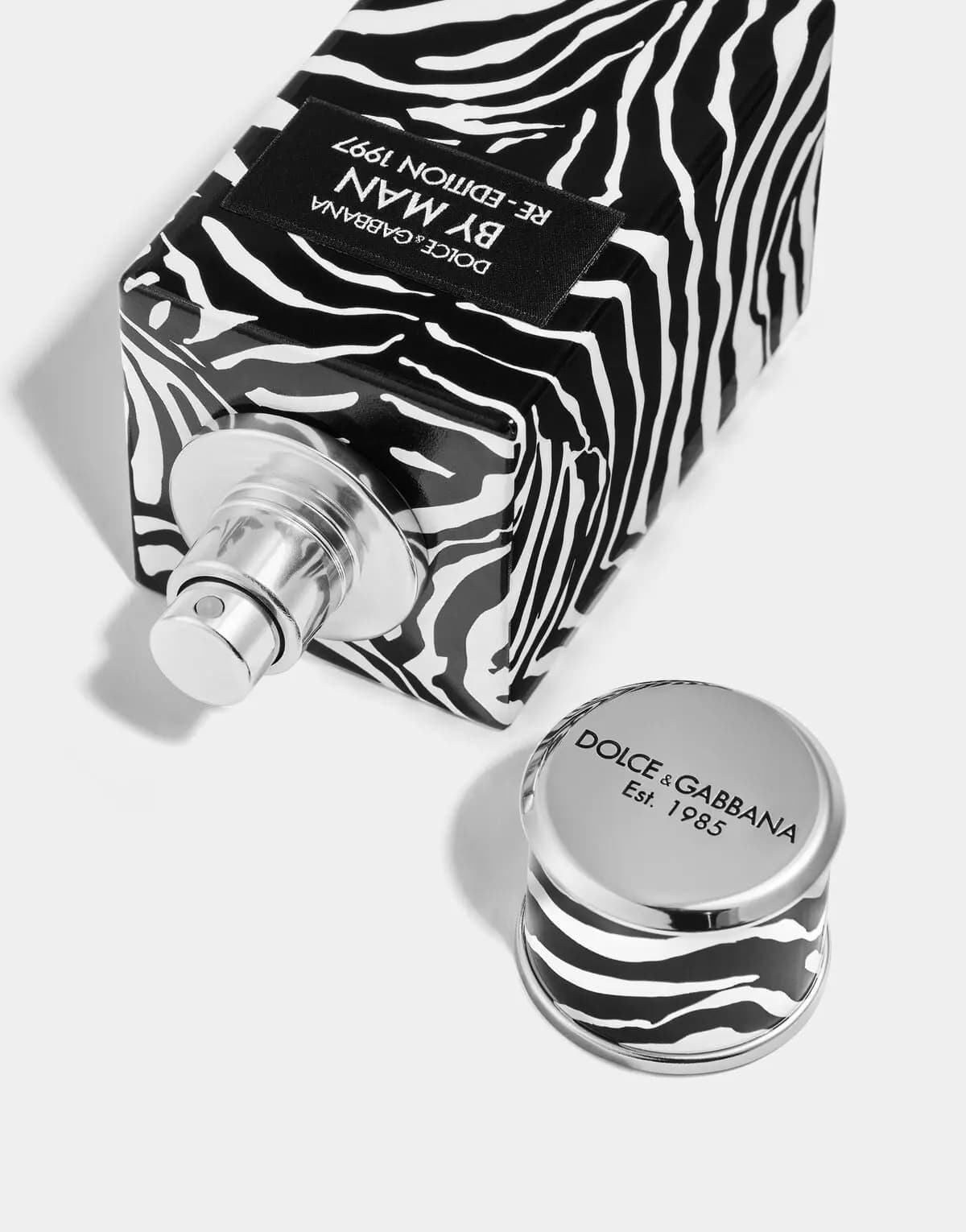 Uncapped zebra-print By Man bottle on its side showing the silver sprayer, beside a cap engraved "Dolce&Gabbana Est. 1985"