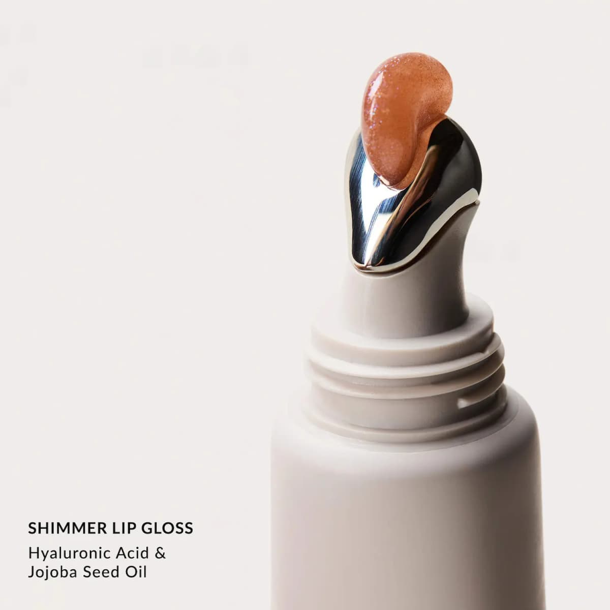 Peach shimmer gloss on the silver metal applicator tip of a Shimmer Lip Gloss tube, noting hyaluronic acid and jojoba