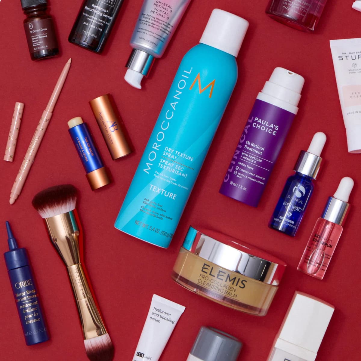 Flat lay on red of Moroccanoil spray, Elemis balm, Paula's Choice retinol, iS Clinical serum, brush and lip products