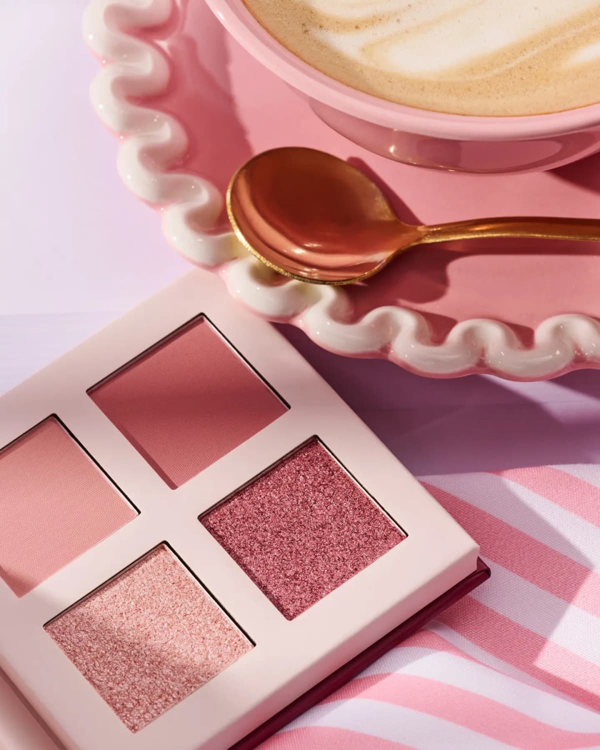 Four-pan eyeshadow palette with matte and shimmer pinks and berry beside a latte on a wavy pink saucer
