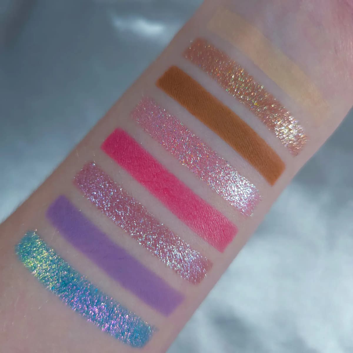 Vertical arm swatches showing matte and shimmer shades from nude to teal-purple multichrome on fair skin