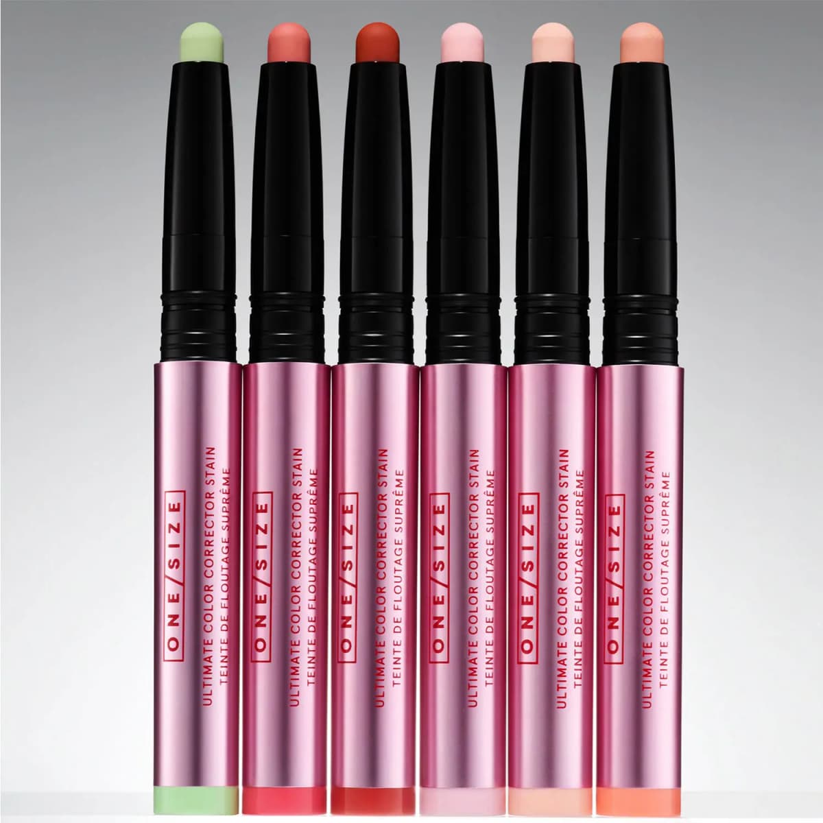All six ONE/SIZE Ultimate Color Corrector Stain sticks standing uncapped in a row: Matcha green and five peach-to-red shades