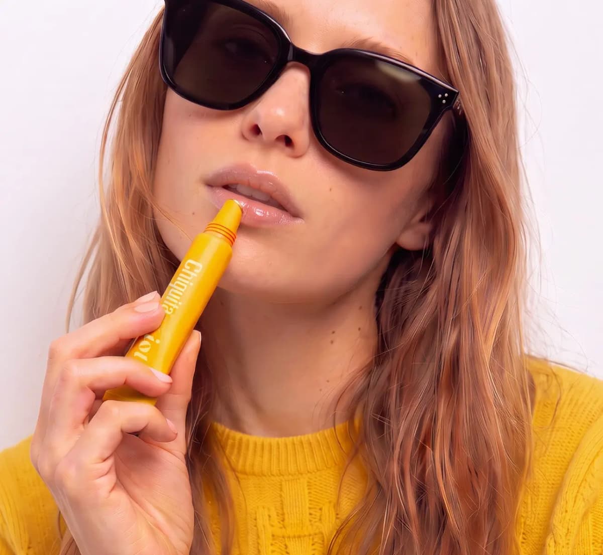 Model in black sunglasses and a yellow sweater dabbing the yellow tube against her shiny lips