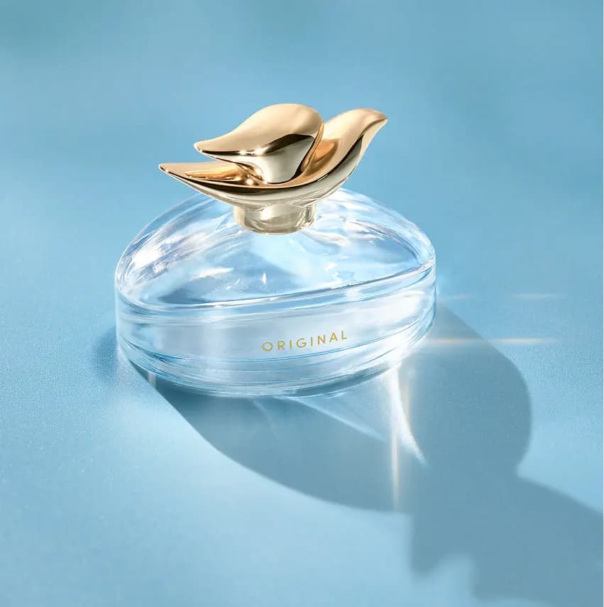 Perfume bottle with gold dove cap on a pale blue surface, casting a long shadow with bright light refractions