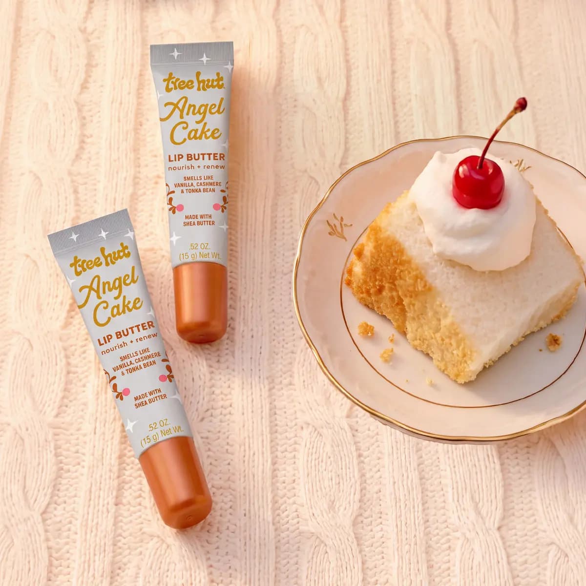 Two Lip Butter tubes on a cream cable knit beside angel cake topped with whipped cream and a cherry