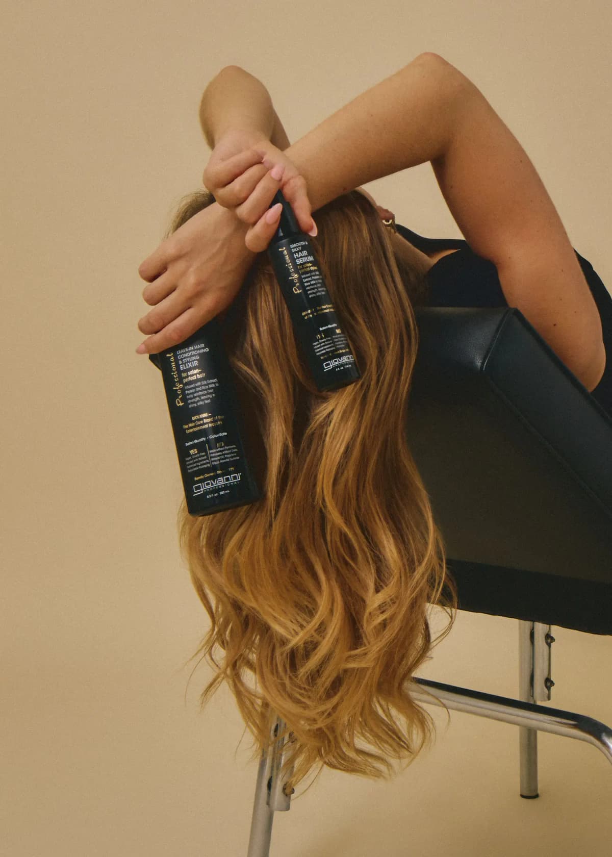Model with long wavy caramel hair hangs head over a black chair, holding the Professional Elixir and Hair Serum.
