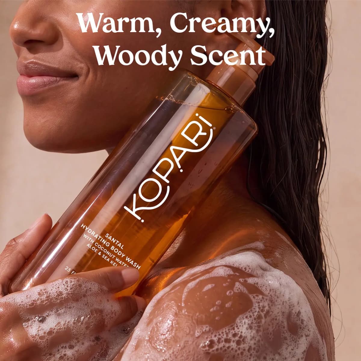 Woman with wet hair holding the amber body wash bottle to her shoulder, text reads Warm, Creamy, Woody Scent.