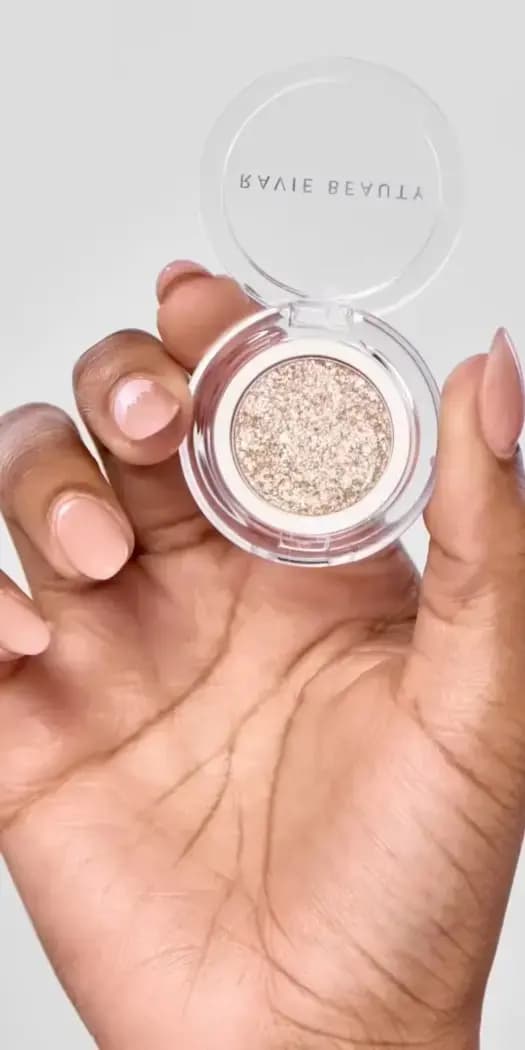 Hand with nude nails holding an open Ravie Beauty compact of champagne-gold sparkling eye topper