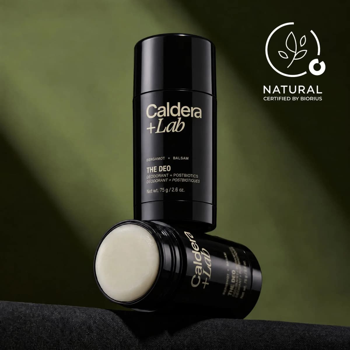 Two black deodorant sticks stacked on dark fabric, one uncapped showing white balm, with a Biorius Natural badge