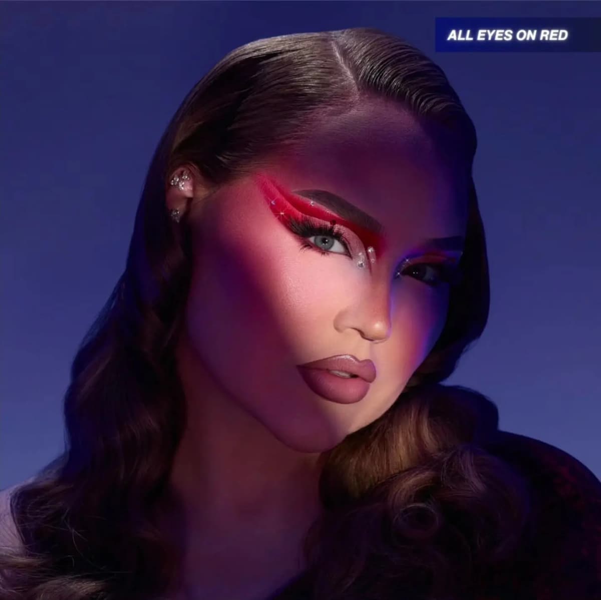 Close-up of NikkieTutorials wearing a red graphic eye look with gem accents and mauve lips, lit dramatically against a deep blue background