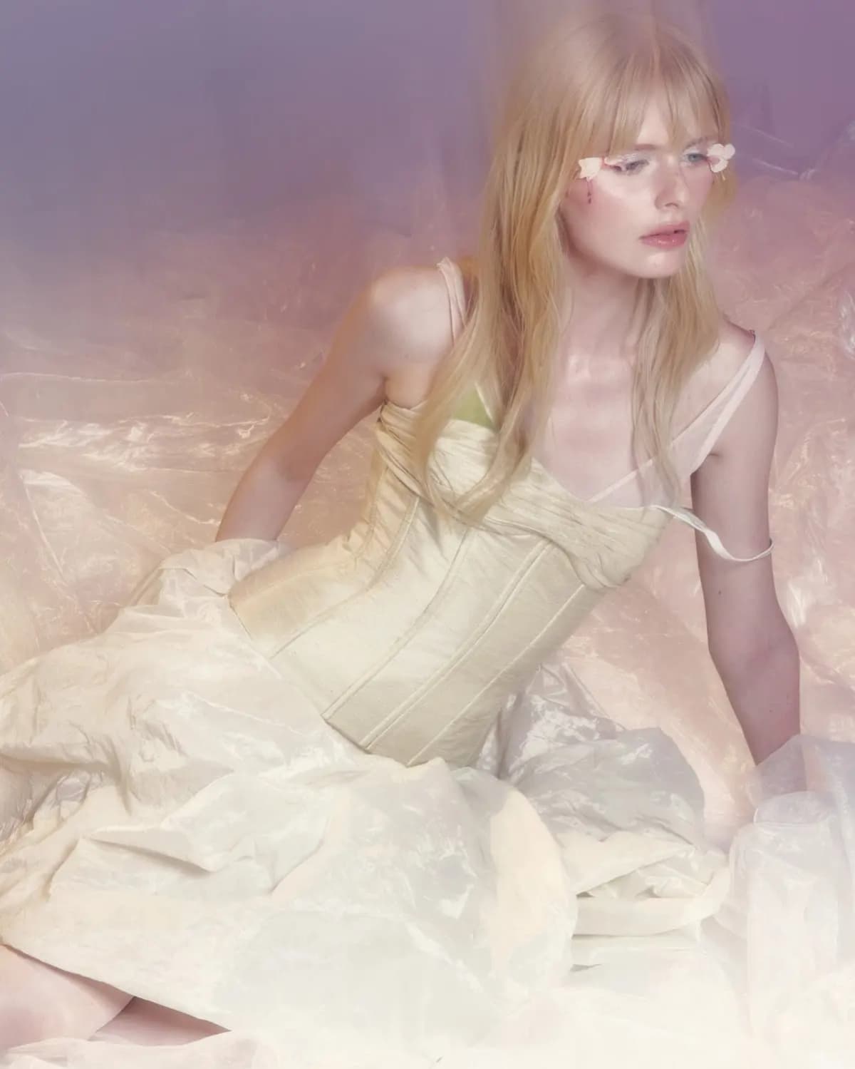Blonde model with flower petals on her eyes in a cream corset gown, seated in pink iridescent sheer fabric