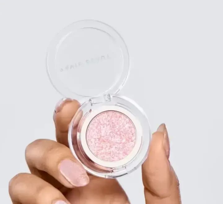 Hand holding an open clear compact of a pink glittery pressed eye topper with Ravie Beauty on the lid