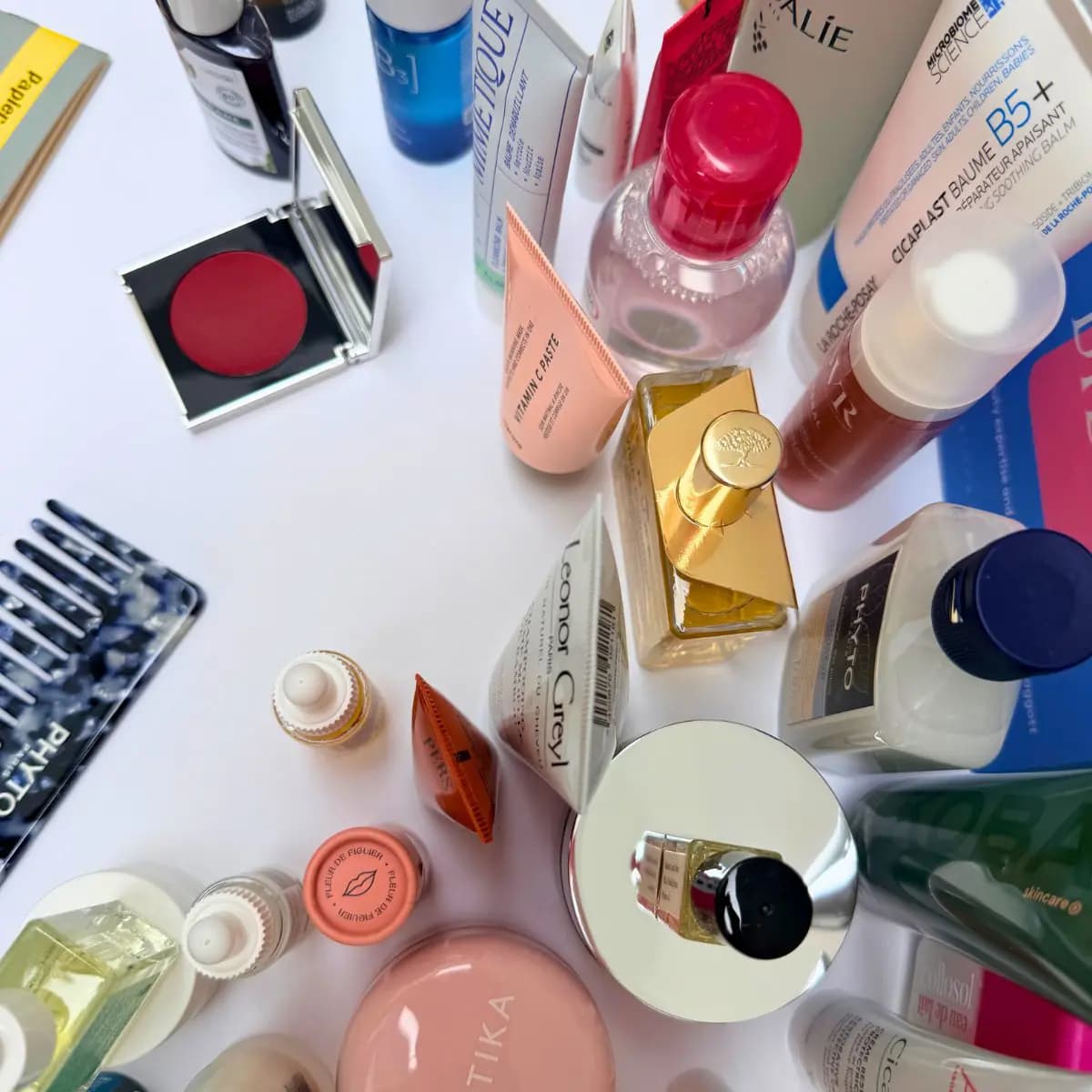 Overhead flat lay of assorted skincare bottles, tubes, a red cream blush compact and a navy patterned Phyto comb