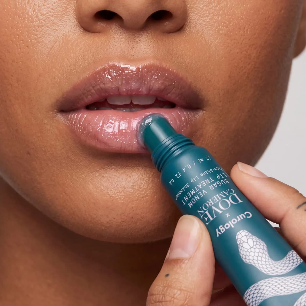 Teal lip tube applicator smoothing glossy clear shimmer onto a model's lower lip