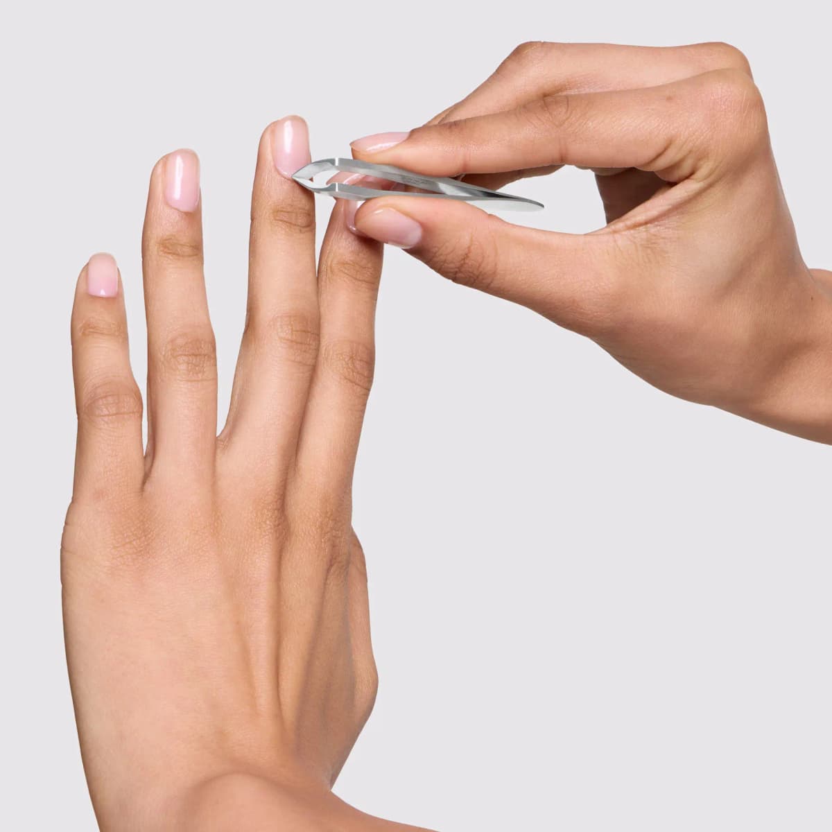 Hand trimming a fingertip cuticle with a stainless steel cuticle nipper against a pale grey background