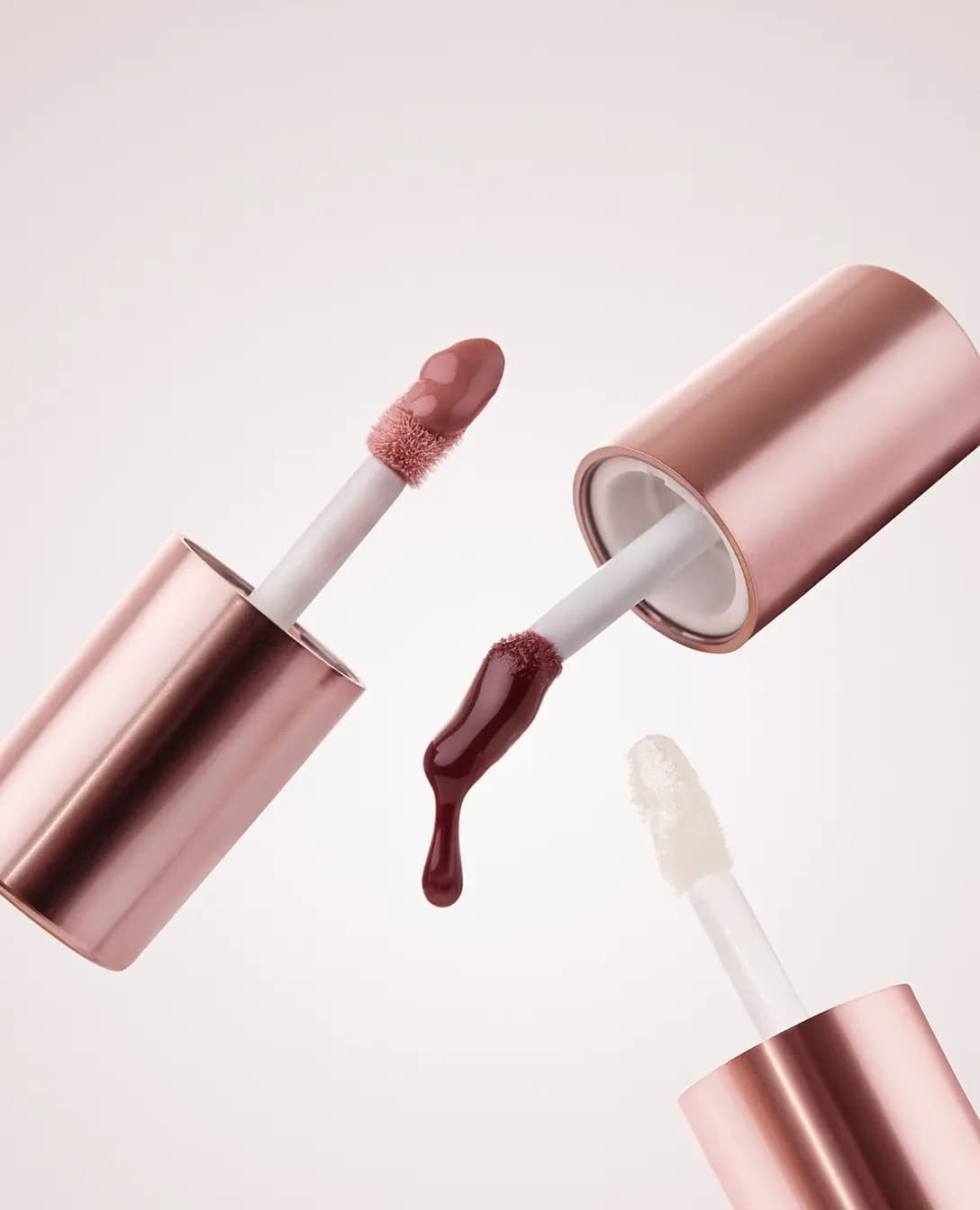 Two rose-gold lip gloss tubes floating open, revealing a plum-brown doe-foot applicator dripping gloss and a clear glittery