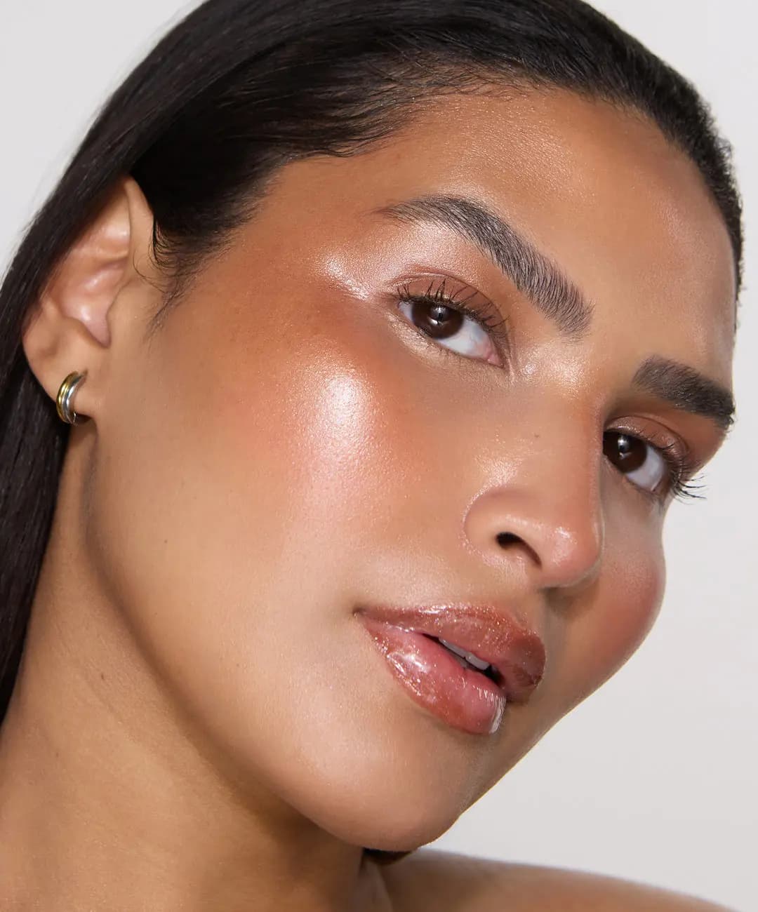 Dark-haired model with intense cheekbone highlight, defined brows and shimmery peach-brown glossy lips