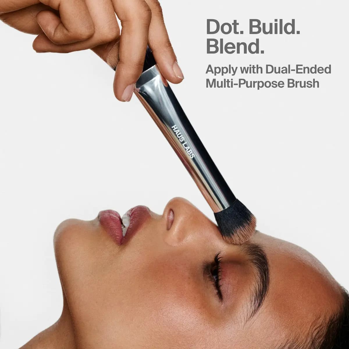 Hand blending bronzer onto a model's temple with a silver Haus Labs brush; text reads 'Dot. Build. Blend.