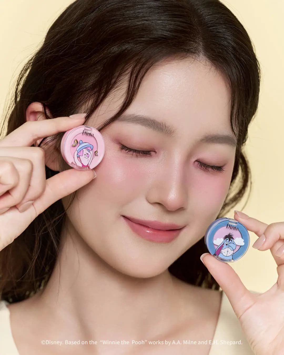 Eyes closed, model with pink lids and cheeks holds round Piglet and Eeyore cheek and eye compacts