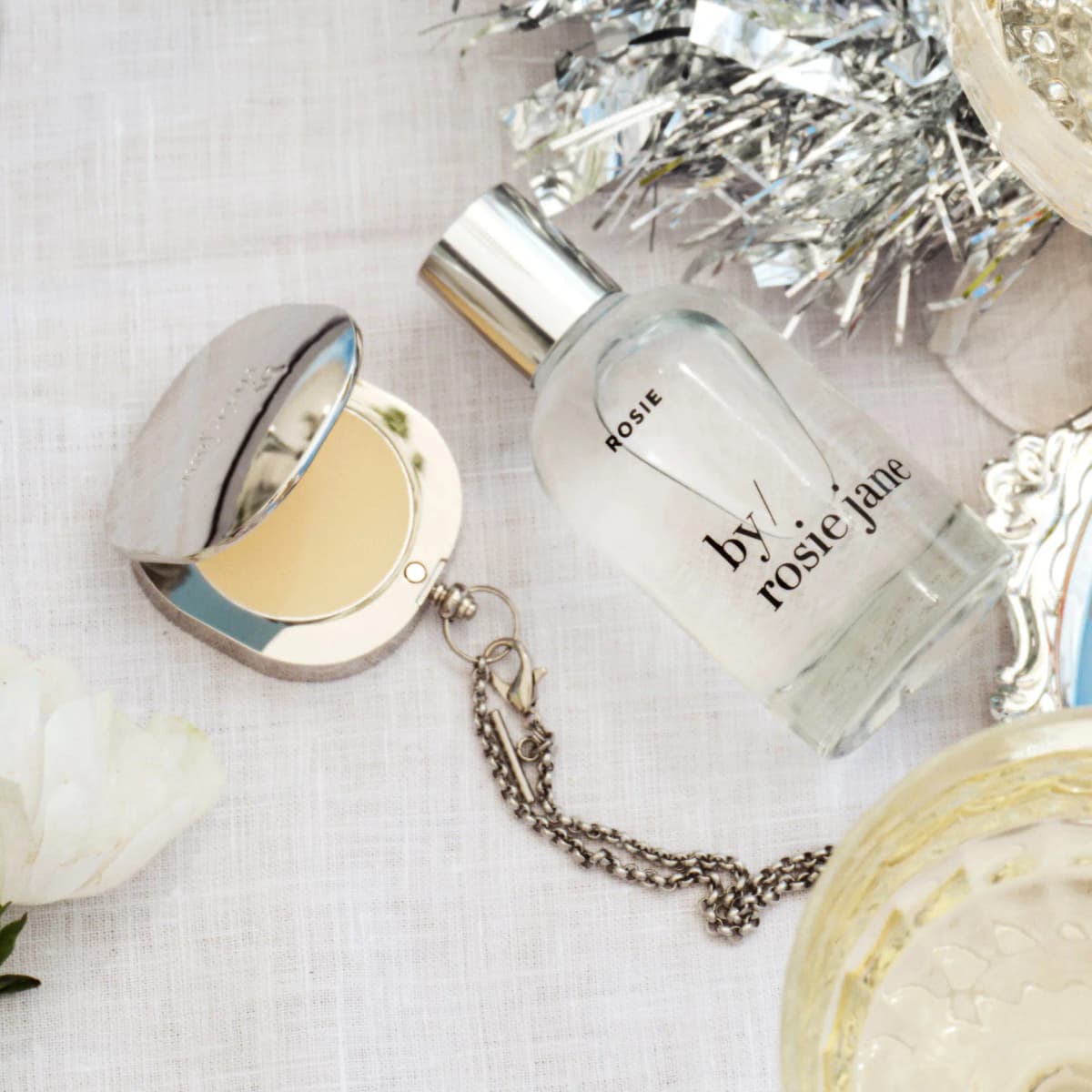 Rosie perfume bottle lying next to an open silver balm compact on a chain, with tinsel and champagne on linen