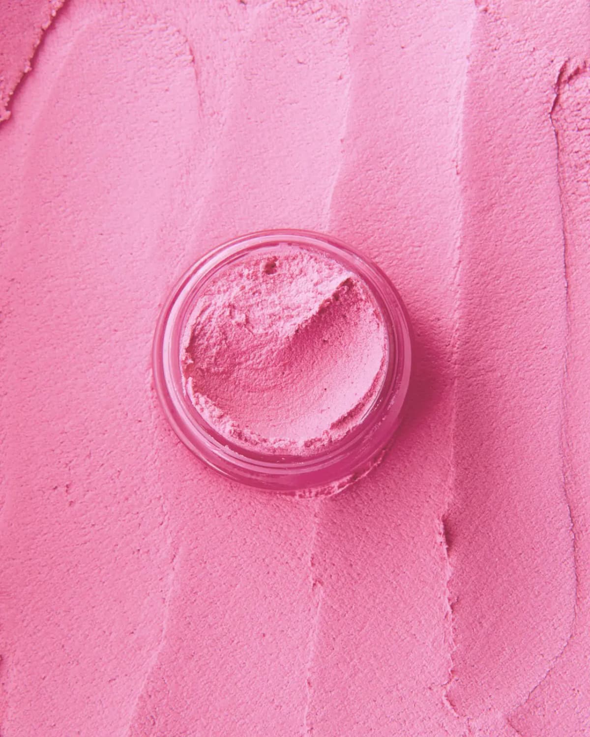 Open jar of pink mousse blush set on a textured backdrop of spread blush in the same shade