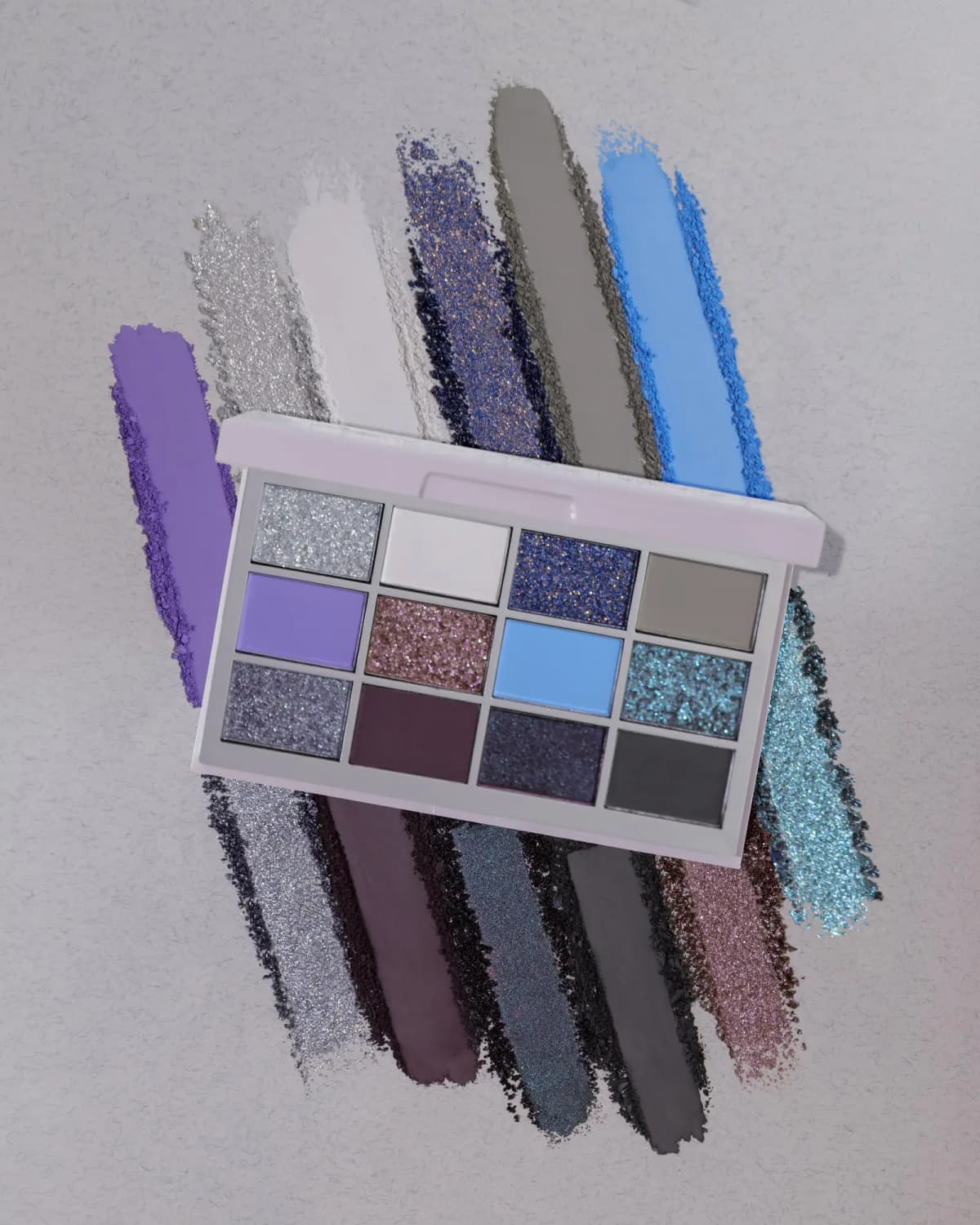 Overhead view of the 12-pan compact laid atop bold diagonal swatches of purple, silver, blue, teal and charcoal shadows.