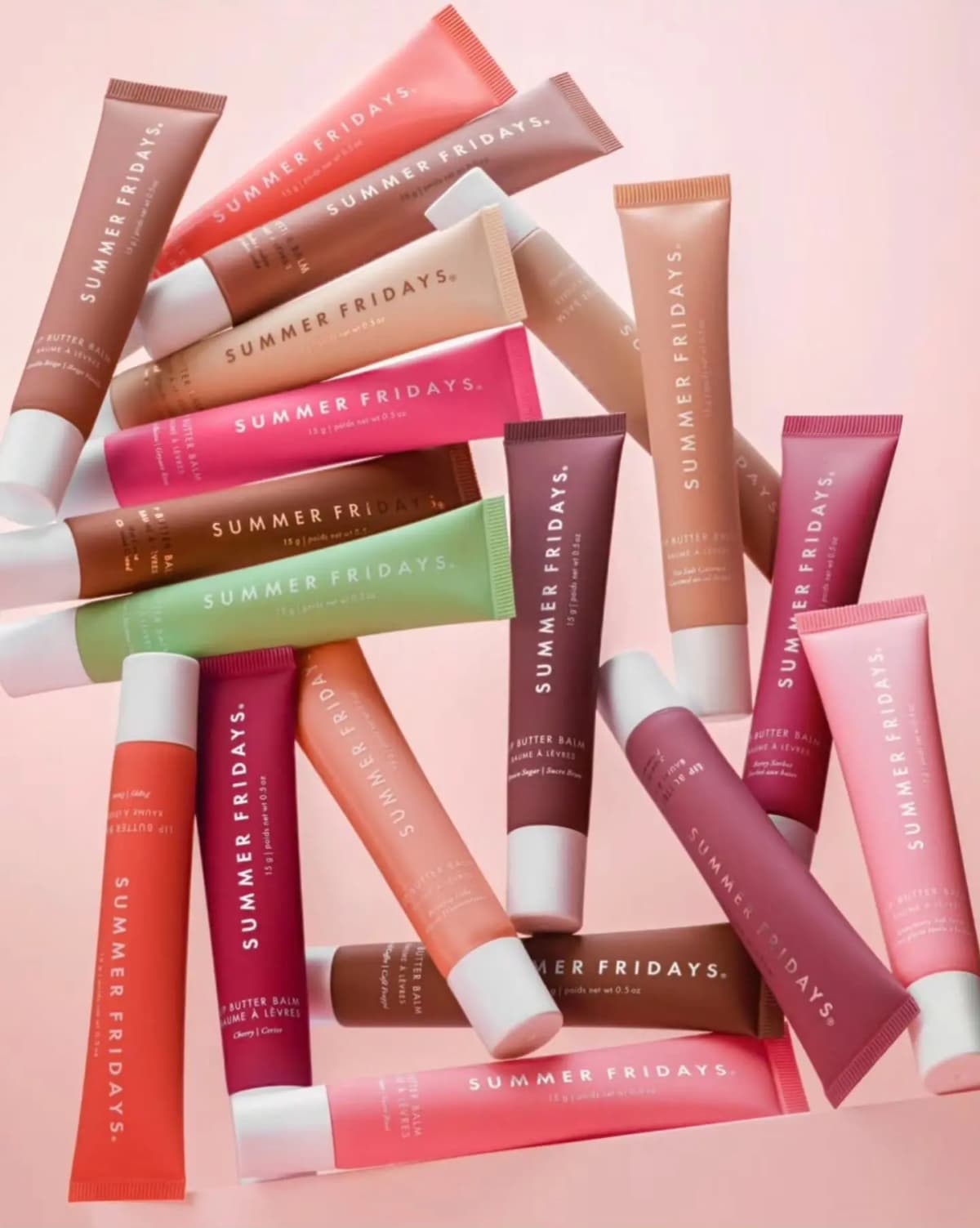 Pile of crisscrossed Lip Butter Balm tubes in pink, red, nude, brown, plum and mint shades on a pale pink background