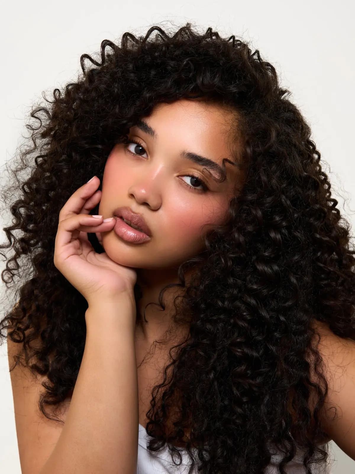 Model with voluminous dark curls touching glossy nude-pink lips, cheeks flushed with soft rosy colour