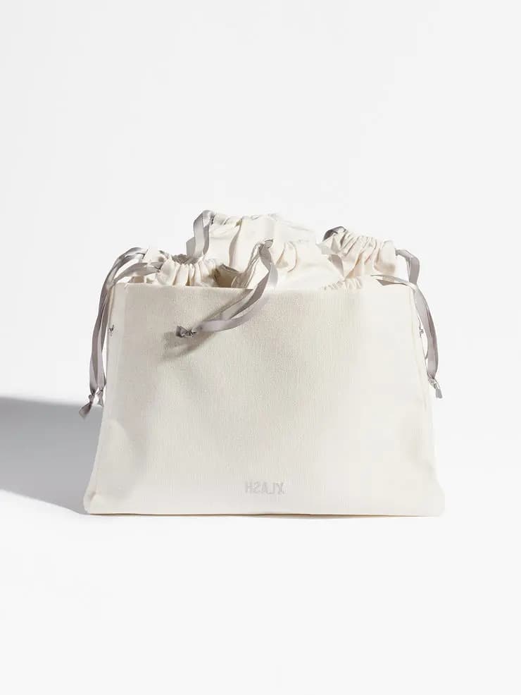 Front view of the cream cotton Xlash bag filled with gathered drawstring pouches tied with grey ribbons