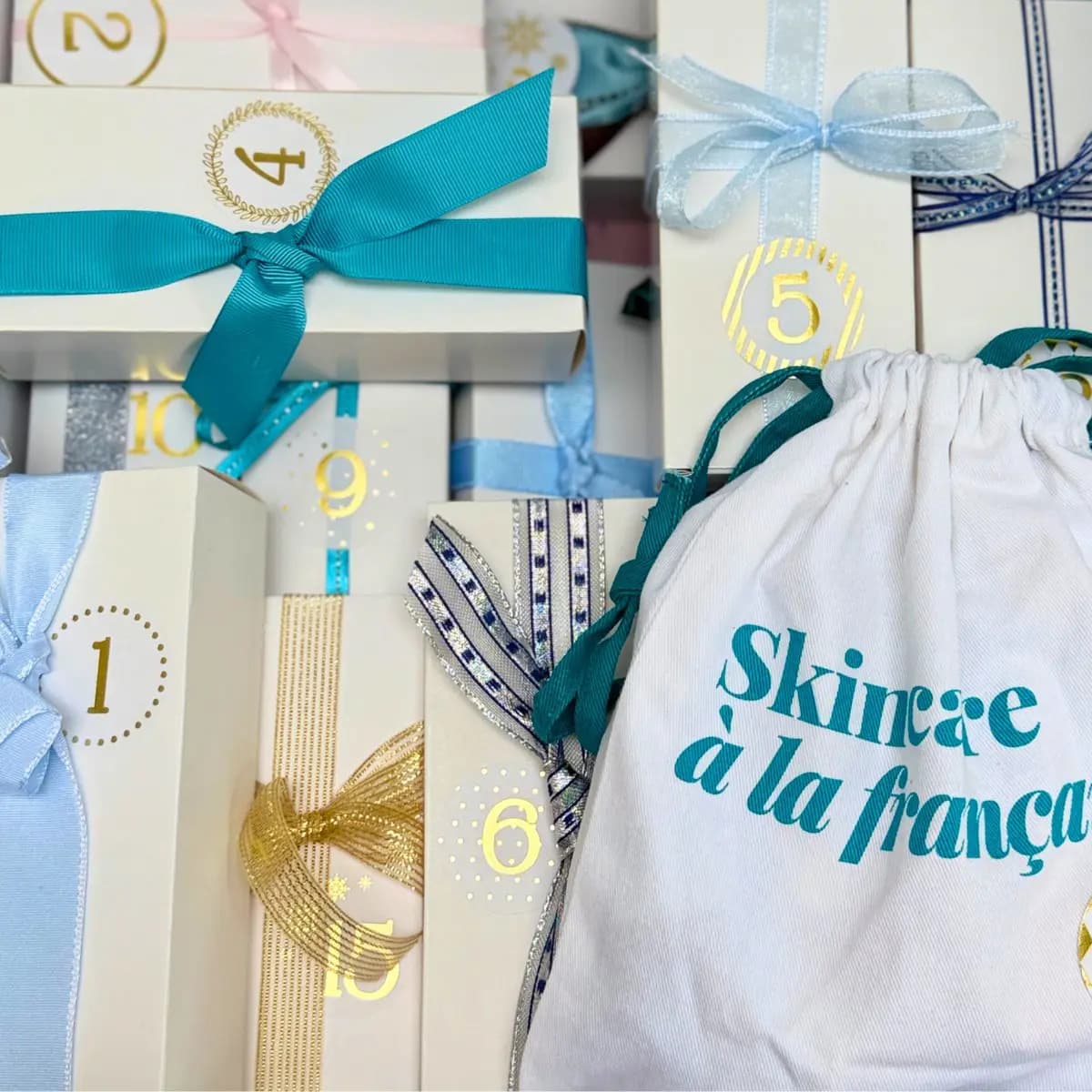Close-up of numbered advent calendar gift boxes tied with teal, blue and gold ribbons beside a Skincare à la française pouch