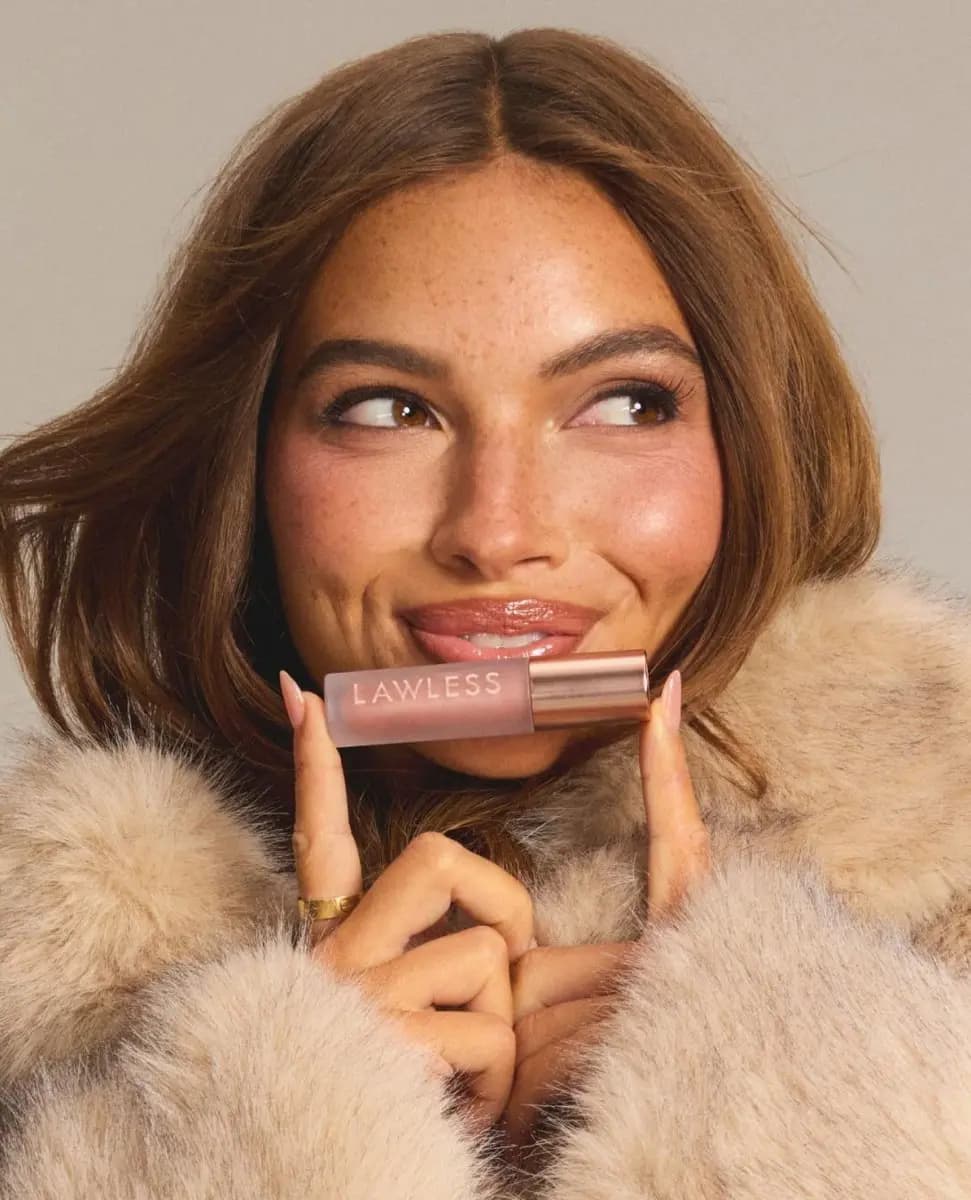A smiling model in a fur coat holding a Lawless lip-plumping gloss tube up to her glossy lips