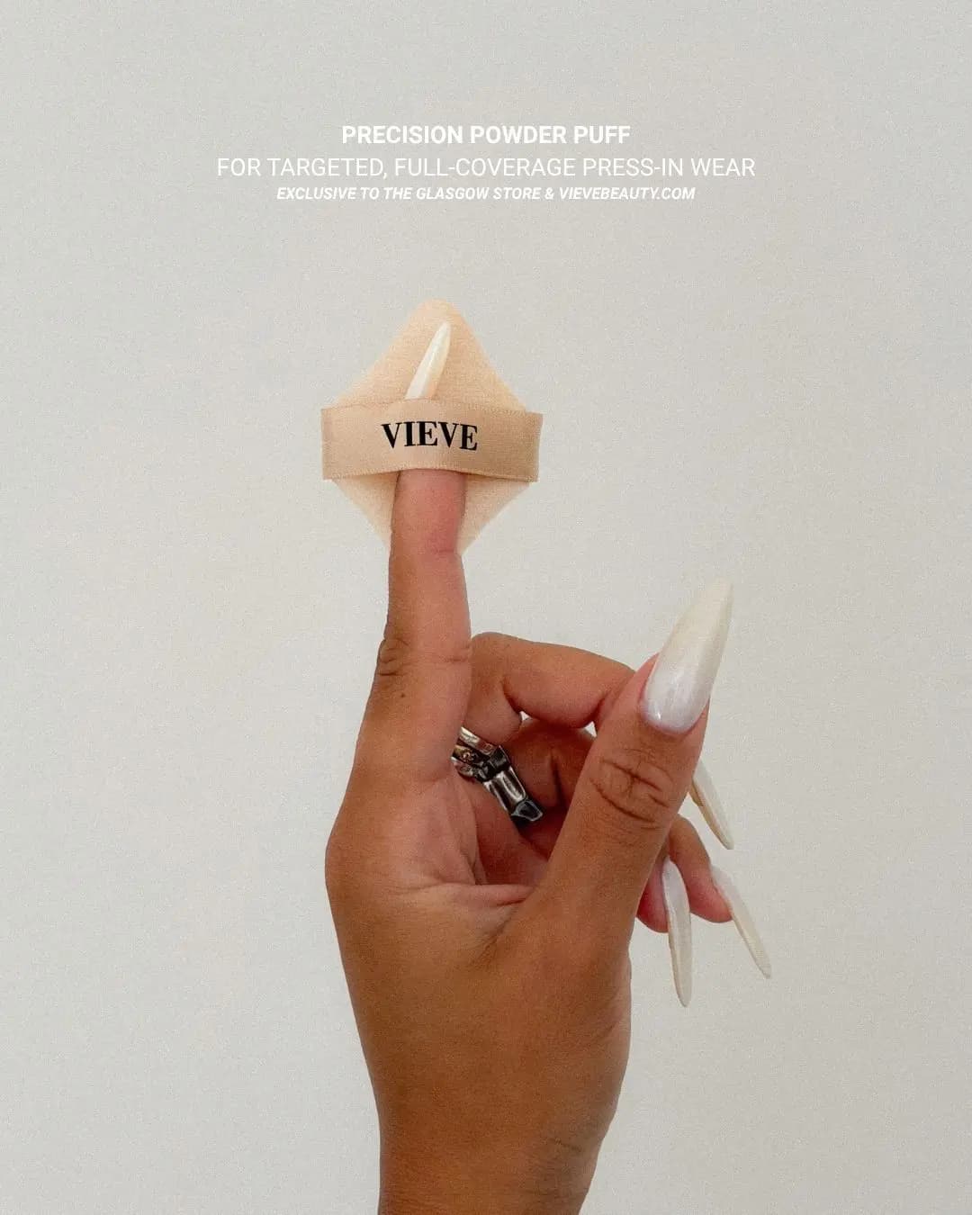 Hand with long pearly nails holding the beige VIEVE Precision Powder Puff on one finger against a pale grey wall