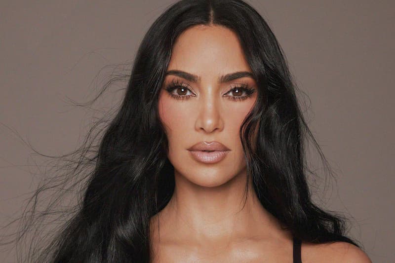 Close-up portrait of Kim Kardashian with wavy black hair, defined brows and glossy nude lips against a taupe backdrop.