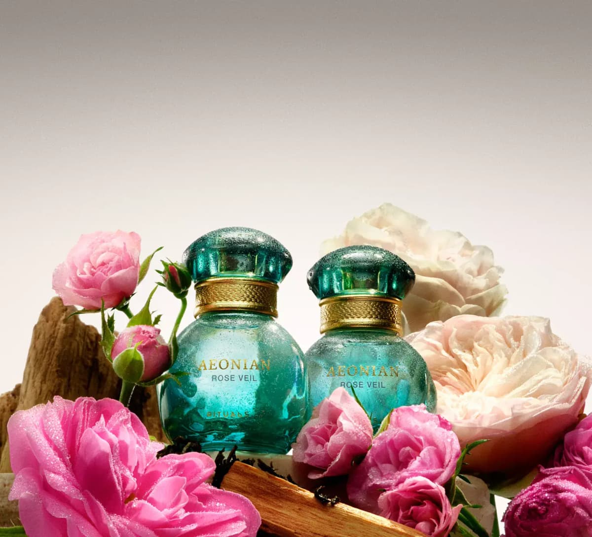 Two aqua Rituals Aeonian Rose Veil bottles nestled among dewy pink and pale peach roses with pale wood