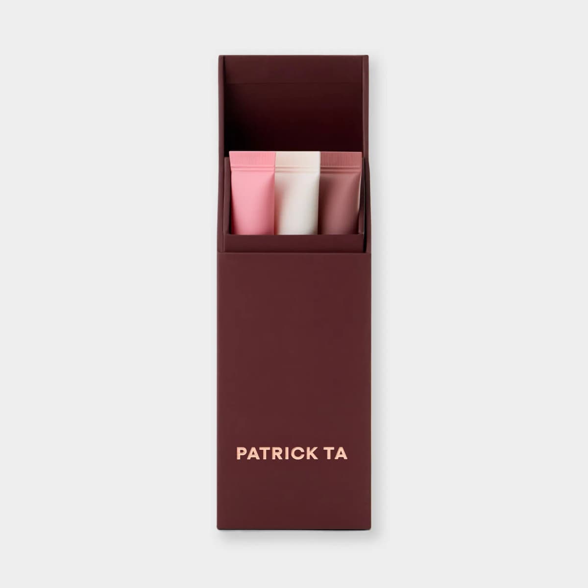 Burgundy box opened to show three lip balm tubes in pink, creamy white and mauve, with rose-gold Patrick Ta logo