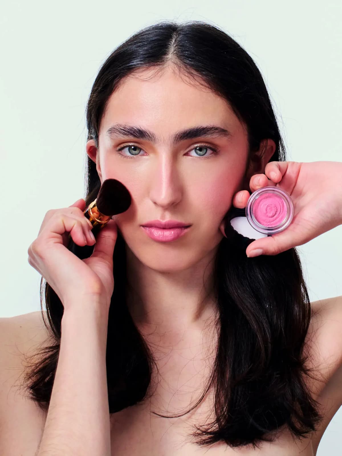 Model sweeps blush onto her cheek with a gold-handled brush while holding an open pink mousse blush jar