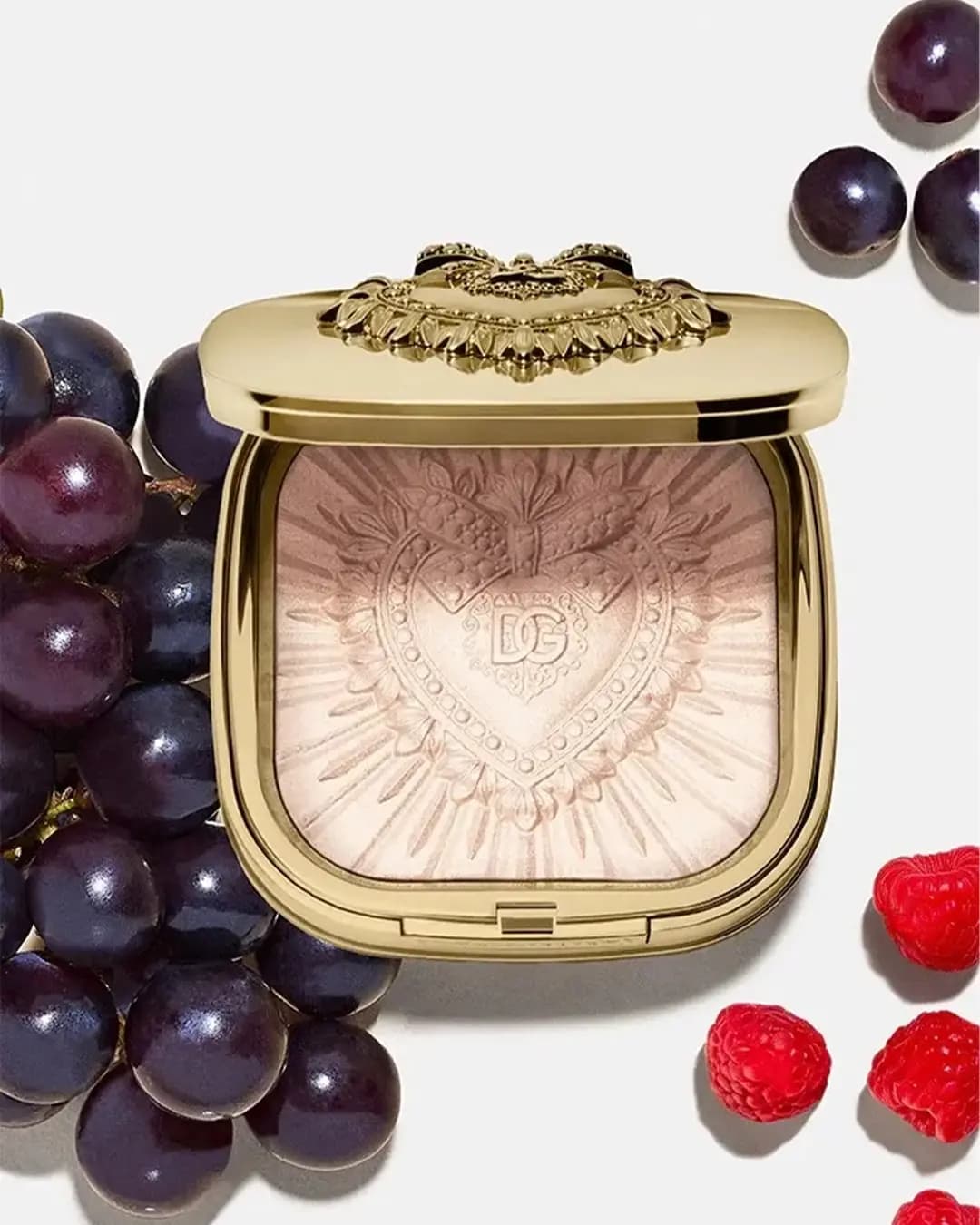Open gold compact with a soft rosy-beige glow powder, surrounded by dark grapes and raspberries
