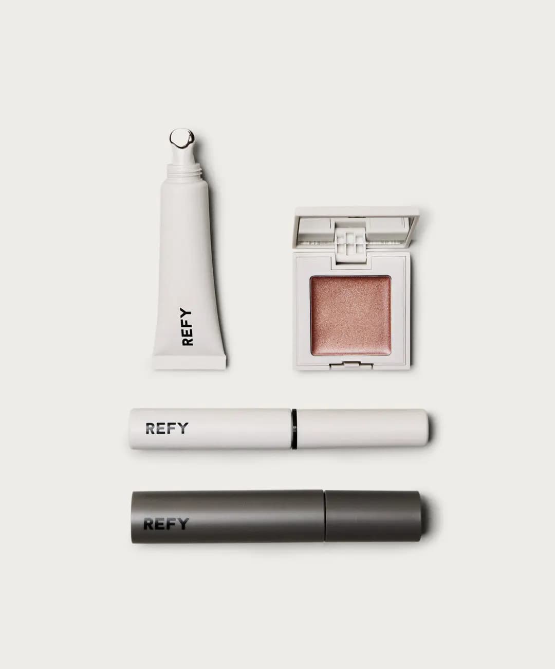 Flat lay of white lip gloss tube, open rose-gold cream shimmer compact, white tube and grey mascara