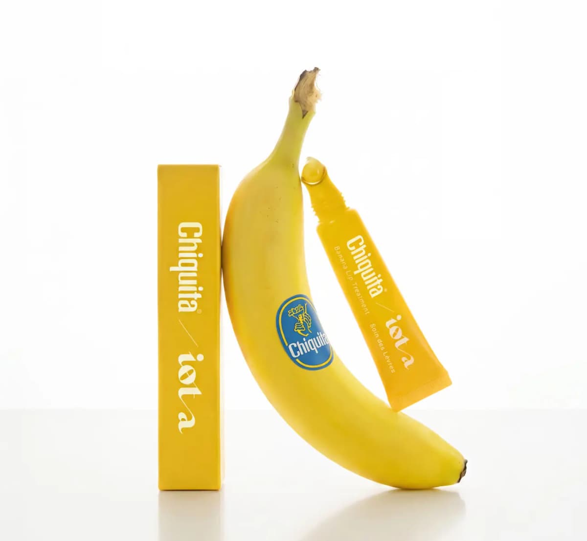 Iota / Chiquita Banana Lip Treatment yellow tube and box propped against a whole banana with a blue Chiquita sticker