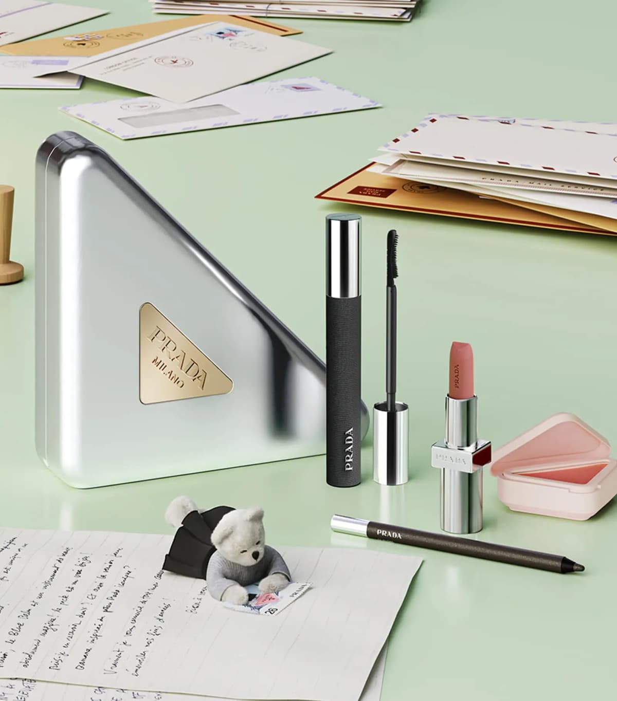 Silver Prada Milano triangle case, mascara, lipstick, pink blush compact and eyeliner on a mint desk with letters